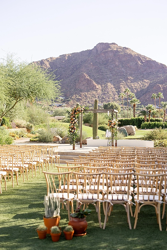 Outdoor ceremony setup with a wood wedding arch and asymmetrical florals, potted cacti in terracotta pots, mountain backdrop under clear sky
