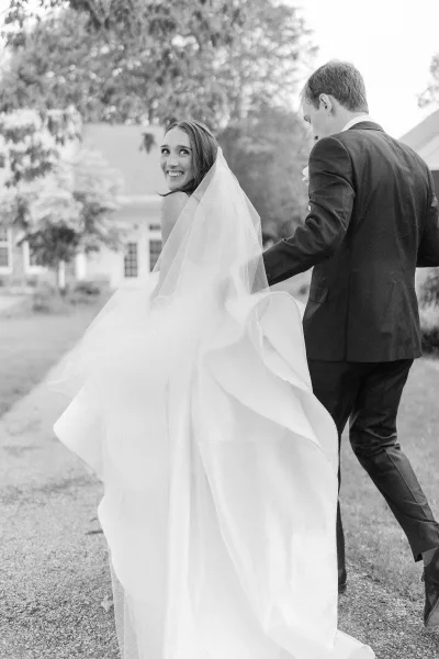 Couple portrait of bride looking back as newlyweds walk away, her long veil and strapless gown trailing on a gravel path by trees and lawn