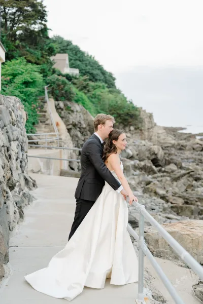 Couple portrait of bride and groom embrace on a coastal walkway, her strapless wedding dress and his tuxedo by a railing above rocky ocean shore