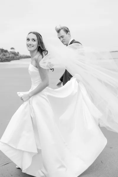 Couple portrait on a beach wedding portrait, bride and groom walking on sand as her long veil blows by the ocean under cloudy skies