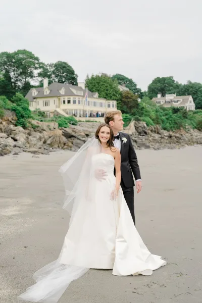 Couple portrait of bride in strapless wedding dress with cathedral veil embracing groom in black tuxedo on a rocky beach under overcast sky