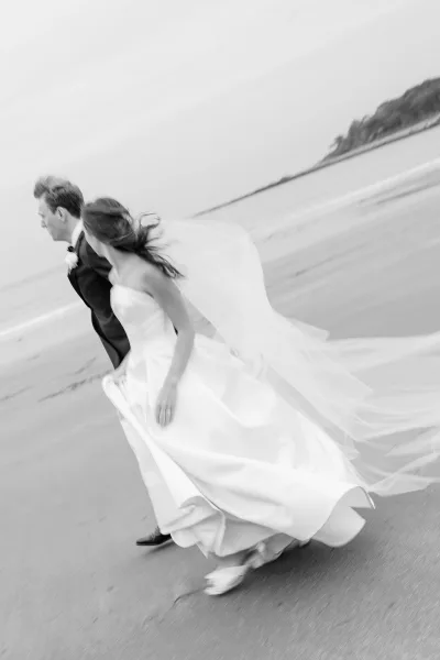 Wedding couple portrait of bride and groom running on the beach, her long veil blowing in the wind with ocean and cliffs behind them