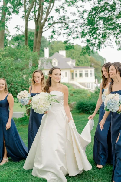 Bride with bridesmaids in navy walking together on a green lawn, the bride in a strapless satin dress holding a white rose bouquet near trees and a house