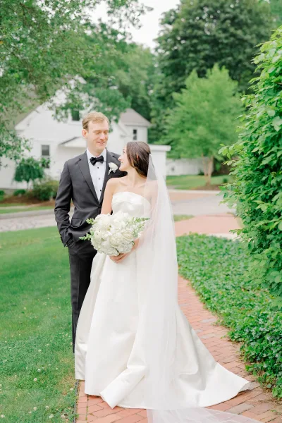 Couple portrait of bride holding a white rose bouquet and groom in black tuxedo, facing each other on a garden lawn by a brick walkway