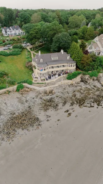 Wedding venue exterior, coastal wedding venue with wraparound porch and large windows overlooking a rocky shoreline, sandy beach, and ocean water