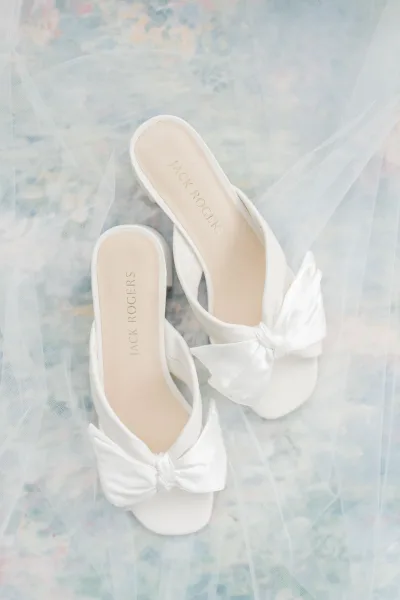 Bridal shoes with white wedding heels featuring satin bow details, styled beside a tulle veil on a pastel patterned flat lay surface