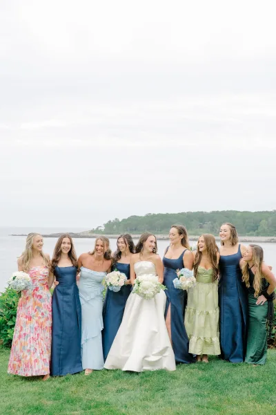 Bridesmaids portrait with bride centered, holding hydrangea bouquets and wearing navy dresses, on an oceanfront lawn by rocky shore