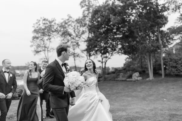 Wedding recessional as bride and groom walking hand in hand, bouquet in his grasp, across a waterfront lawn under cloudy sky with party behind