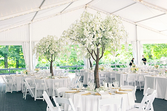 Reception tablescape in a wedding tent reception with round white tables, woven gold chargers, tall flowering branches, candles, and string lights overhead