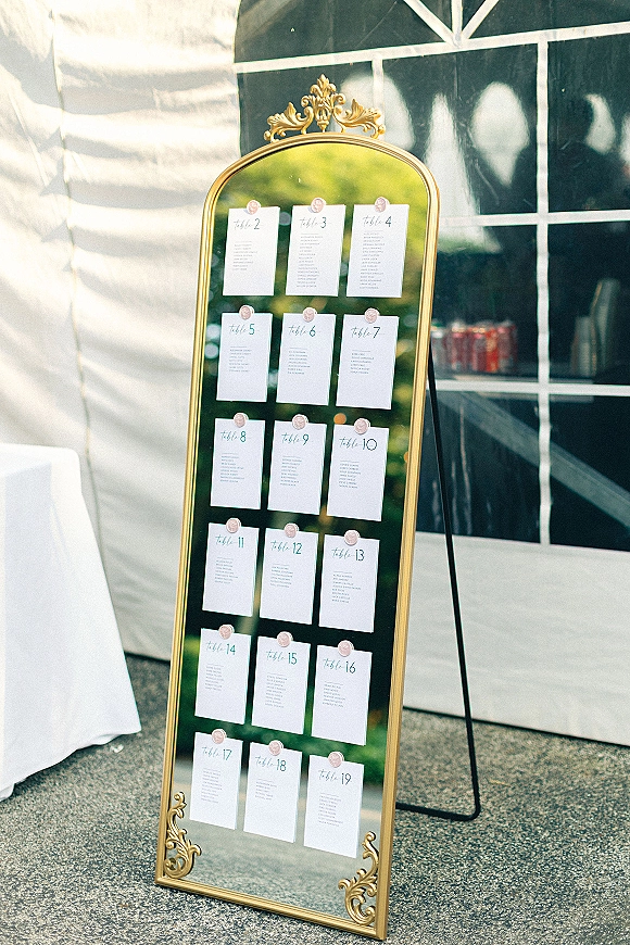 Wedding seating chart on an ornate gold mirror with escort cards and wax seals, displayed on an easel by a white tent wall
