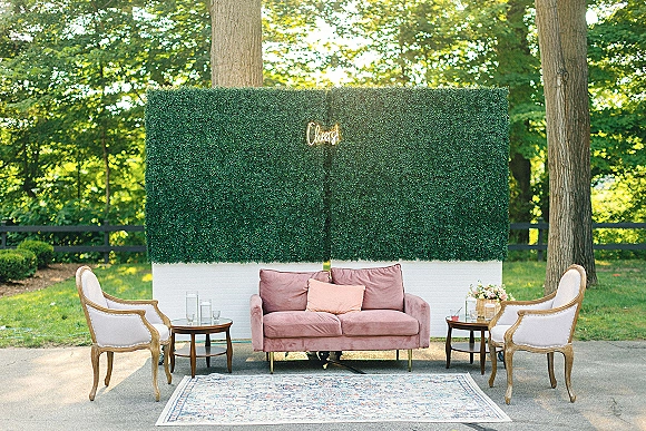 Wedding lounge seating with a pink velvet sofa, upholstered armchairs, pillows, and side tables set before a hedge wall with neon sign on a lawn patio