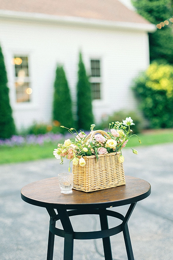 Wedding floral centerpiece in a wicker basket with pastel blooms and greenery on a wooden cocktail table beside a glass votive candle outdoors