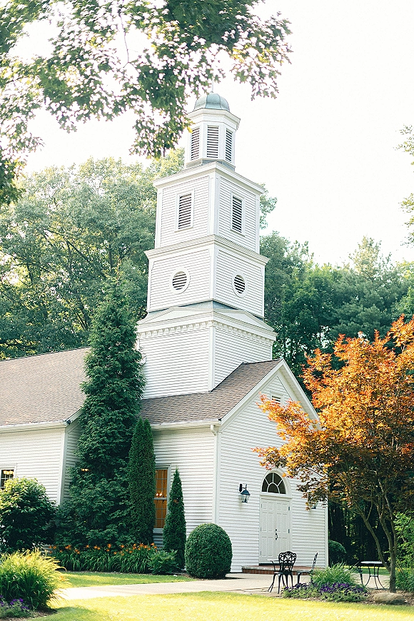 Wedding chapel exterior with white chapel exterior siding, steeple tower and arched doorway, plus bistro patio seating beside flower beds and trees
