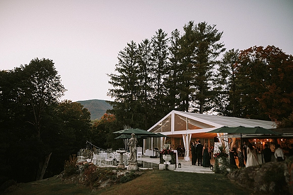 Outdoor wedding reception under a clear top tent with string lights, cocktail tables, umbrellas, florals and lanterns facing mountains