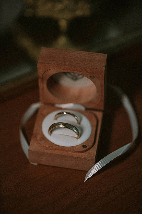 Wedding rings resting in a wood ring box with a satin ribbon on a wood surface, with soft greenery bokeh in the background