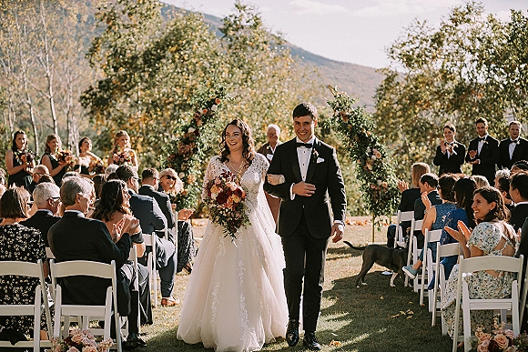 Wedding recessional as bride and groom walk the aisle, bride holding a bouquet under a greenery arch with guests and mountains behind