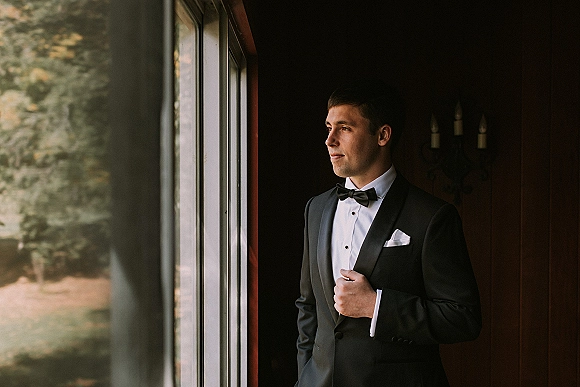 Groom portrait in a classic black tuxedo and bow tie, looking out a window beside a wood-paneled wall with trees beyond