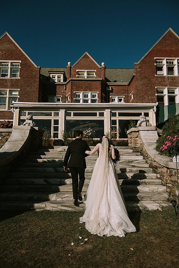 Couple portrait of newlyweds walking up stone steps, bride in lace gown with cathedral veil and bouquet beside groom in tuxedo