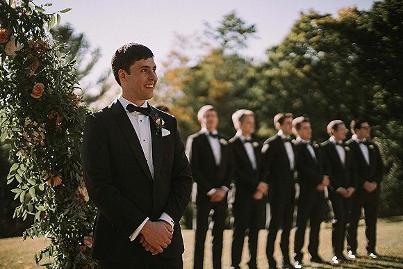 Groom portrait of a black tie groom in a black tuxedo with bow tie and boutonniere, standing by a floral arch in a garden ceremony