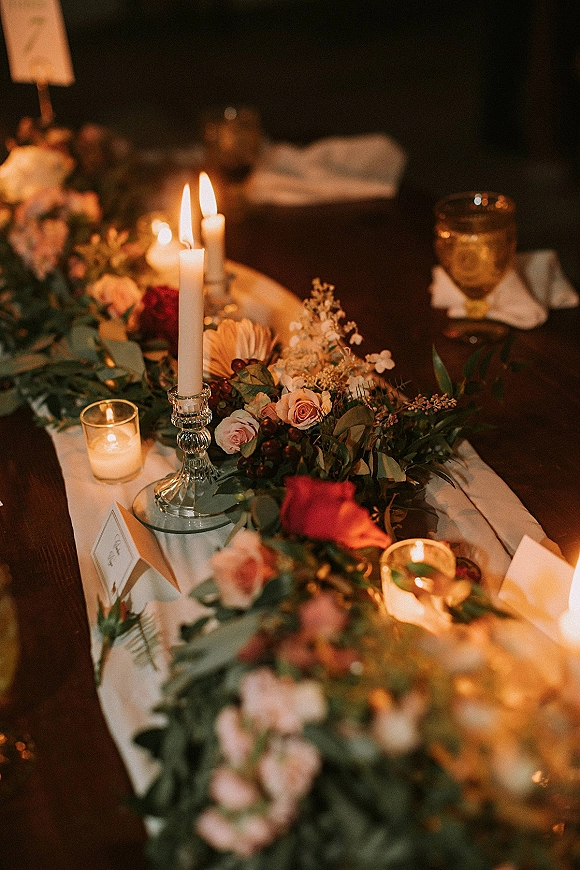 Reception tablescape with wedding table garland of roses and greenery, taper and votive candles glowing on a wood table in low light
