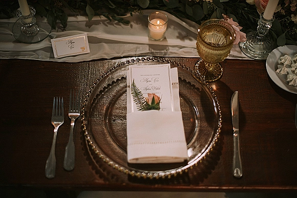 Reception tablescape with a wedding place setting featuring a beaded charger, menu and place cards, amber goblets, and candlelight on a dark wood table