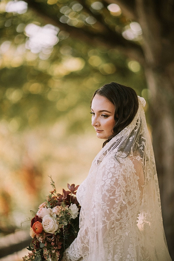 Bridal portrait of a bride in side profile wearing a lace veil, holding a rose and ranunculus bouquet in a sunlit garden with trees