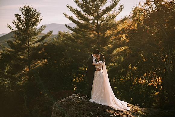 Wedding kiss portrait of bride and groom kissing on a rocky cliff, veil blowing over lace sleeves with pine forest and mountains behind