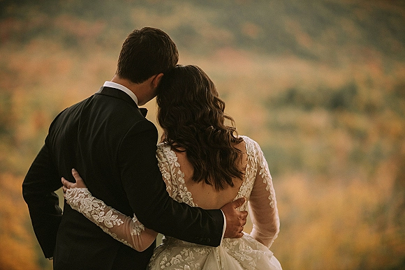 Couple portrait of bride and groom from behind embracing, her long-sleeve lace open-back gown and his tuxedo, facing autumn mountains