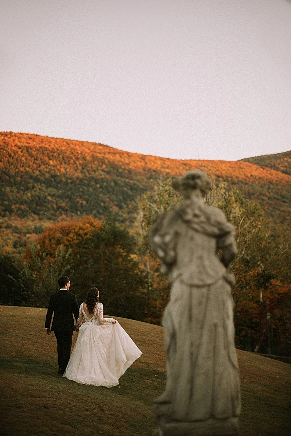 Couple portrait of bride and groom walking away holding hands, bride in lace sleeve dress with long train on a fall mountain hillside.