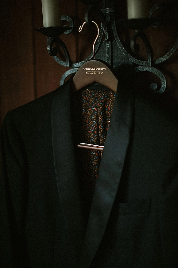 Groom suit hanging on a wooden hanger with metal hook, suit jacket and dress shirt displayed against warm wood paneling