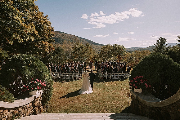Ceremony processional as bride walking down aisle with long train beside groom in suit, guests by white chairs, mountains beyond