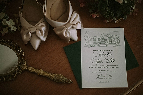 Wedding invitation suite flat lay with green envelope, script cards, blush satin bow heels, vintage mirror, and greenery on wood tabletop