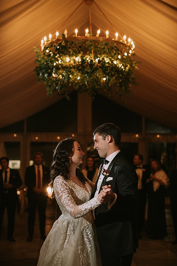 First dance as bride in lace wedding dress and groom in tuxedo sway under a greenery chandelier with candles, guests watching beneath a draped tent ceiling
