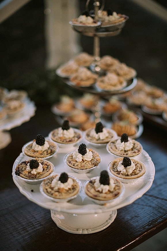 Wedding dessert table with mini pie dessert bar featuring blackberry garnish and whipped cream on tiered stands atop a dark wooden table