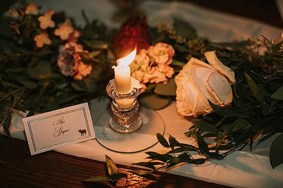 Wedding place setting with wedding place card on a dinner plate, candle in a glass candlestick, and a floral garland on a rustic wood table.