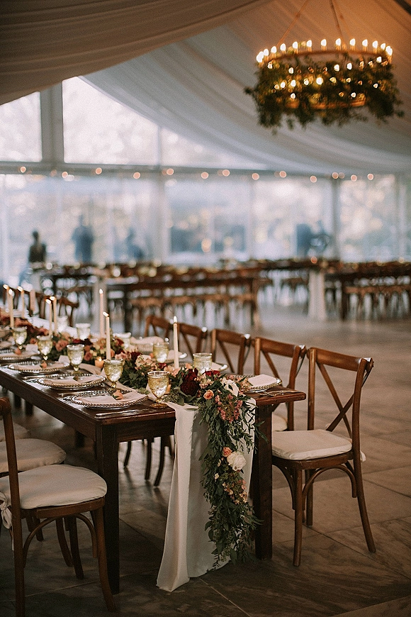 Reception tablescape with a farmhouse head table featuring taper candles and floral garland on a wood table under a draped clear tent with lights