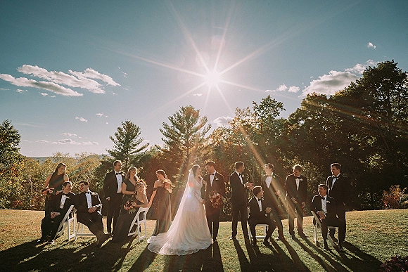 Wedding party portrait of bride and groom with wedding party lined up on a sunny lawn, bridesmaids holding bouquets and groomsmen in tuxedos