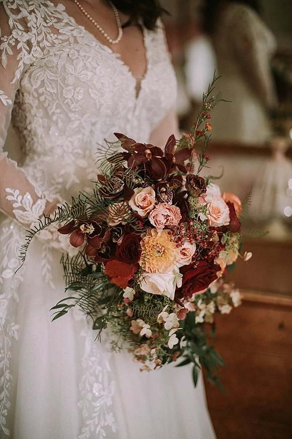 Bridal bouquet in burgundy tones with orchids, roses, dahlias, berries, and greenery held against a lace deep V dress in a softly blurred room