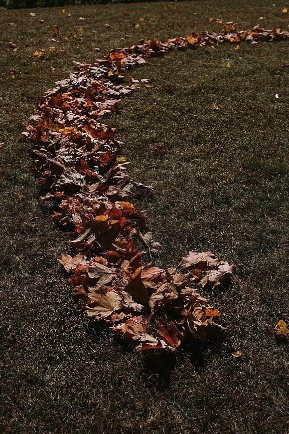 Leaf aisle decor lining an outdoor wedding aisle on a grass lawn, with fallen leaves creating a simple, rustic autumn ceremony path