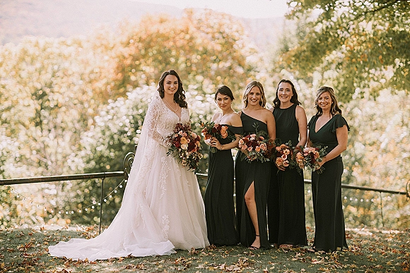 Bride and bridesmaids in black dresses pose with rust and burgundy bouquets under string lights beside autumn trees and fallen leaves