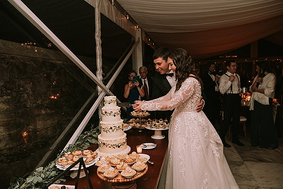 Wedding cake cutting with bride and groom cake cutting at a candlelit dessert table, greenery garland, pastries, and string lights under tent drapery
