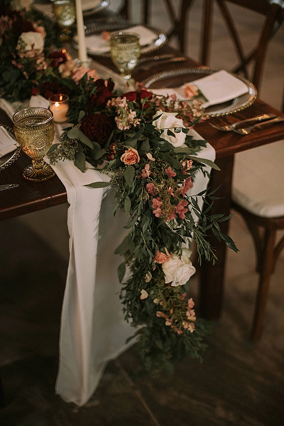 Reception tablescape with wedding table garland of greenery and roses, taper and votive candles, and gold goblets on a rustic wood table
