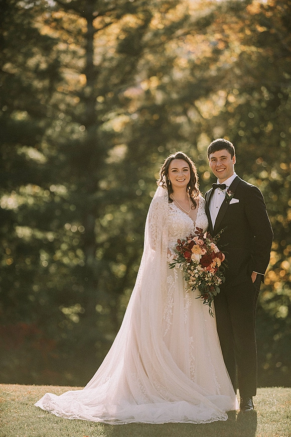 Couple portrait of bride and groom outdoors, bride holding bouquet with a lace veil beside groom in tuxedo, sunlit trees behind