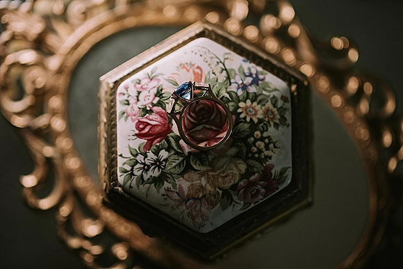 Engagement ring with a blue sapphire gemstone in a floral ring box, styled beside an ornate gold frame on a dark surface