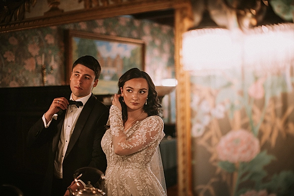 Couple portrait in an ornate mirror, bride in long-sleeve lace dress and veil as groom adjusts bow tie in a vintage room