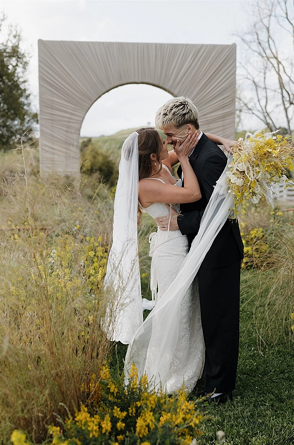 Wedding couple portrait of bride and groom embracing, her veil blowing as she holds his face before a fabric arch in a wildflower meadow