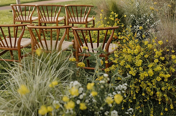 Ceremony seating with wooden outdoor ceremony chairs and woven seats, lined with yellow and white flowers and grasses on a garden lawn