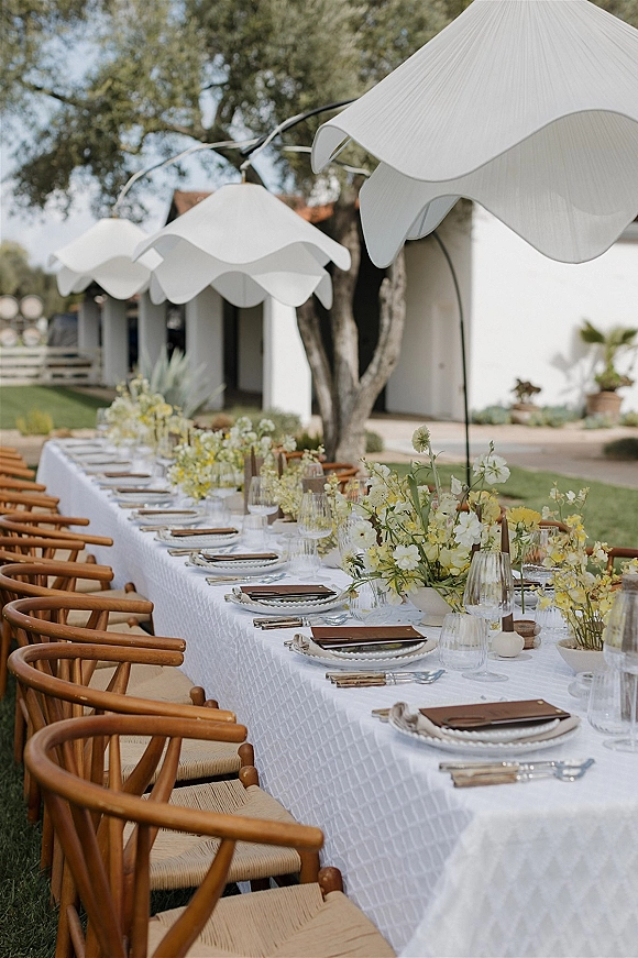 Reception tablescape with a long banquet table setup on a lawn, white tablecloth, wood chairs, taper candles, bud vases, and white umbrellas