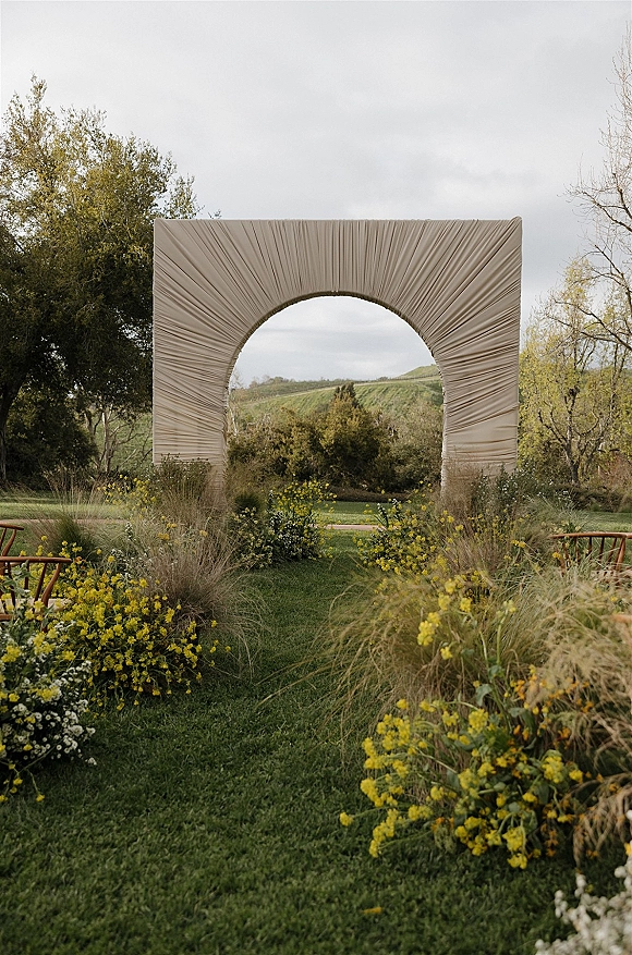 Ceremony backdrop with fabric draped arch and yellow and white florals, framed by wooden chairs on a grassy lawn with hills beyond