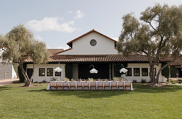 Outdoor reception tablescape with a long banquet table, white tablecloth, floral centerpieces and taper candles in a Spanish courtyard with pendant lights